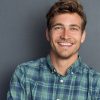 Young handsome man leaning against grey wall with arms crossed. Cheerful man laughing and looking at camera with a big grin. Portrait of a happy young man standing with crossed arms over grey background.